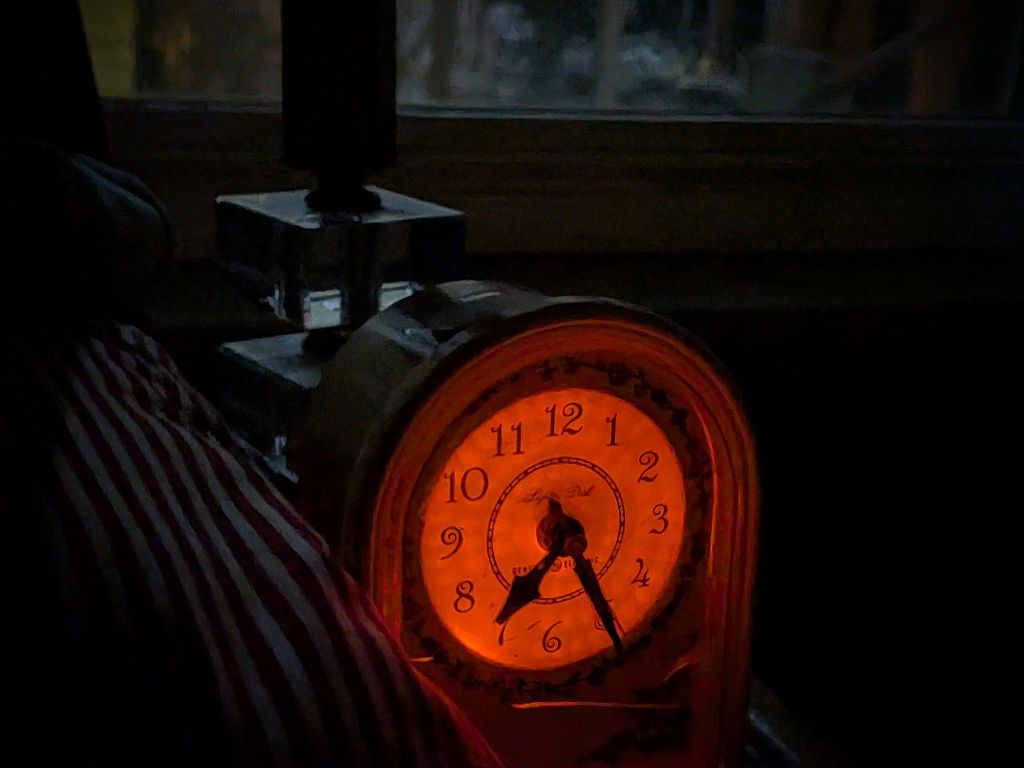 Picture of old-fashion alarm clock, the plug-in kind, with illuminated face. In front of window illuminated by moonlight