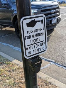 A "Beg button" on a post at a pedestrian crossing. Large metallic button with an arroiw, and sign reads: "Push button for warning lights. Wait for gap in traffic." A Ford F-150 truck is passing by just off the curb.